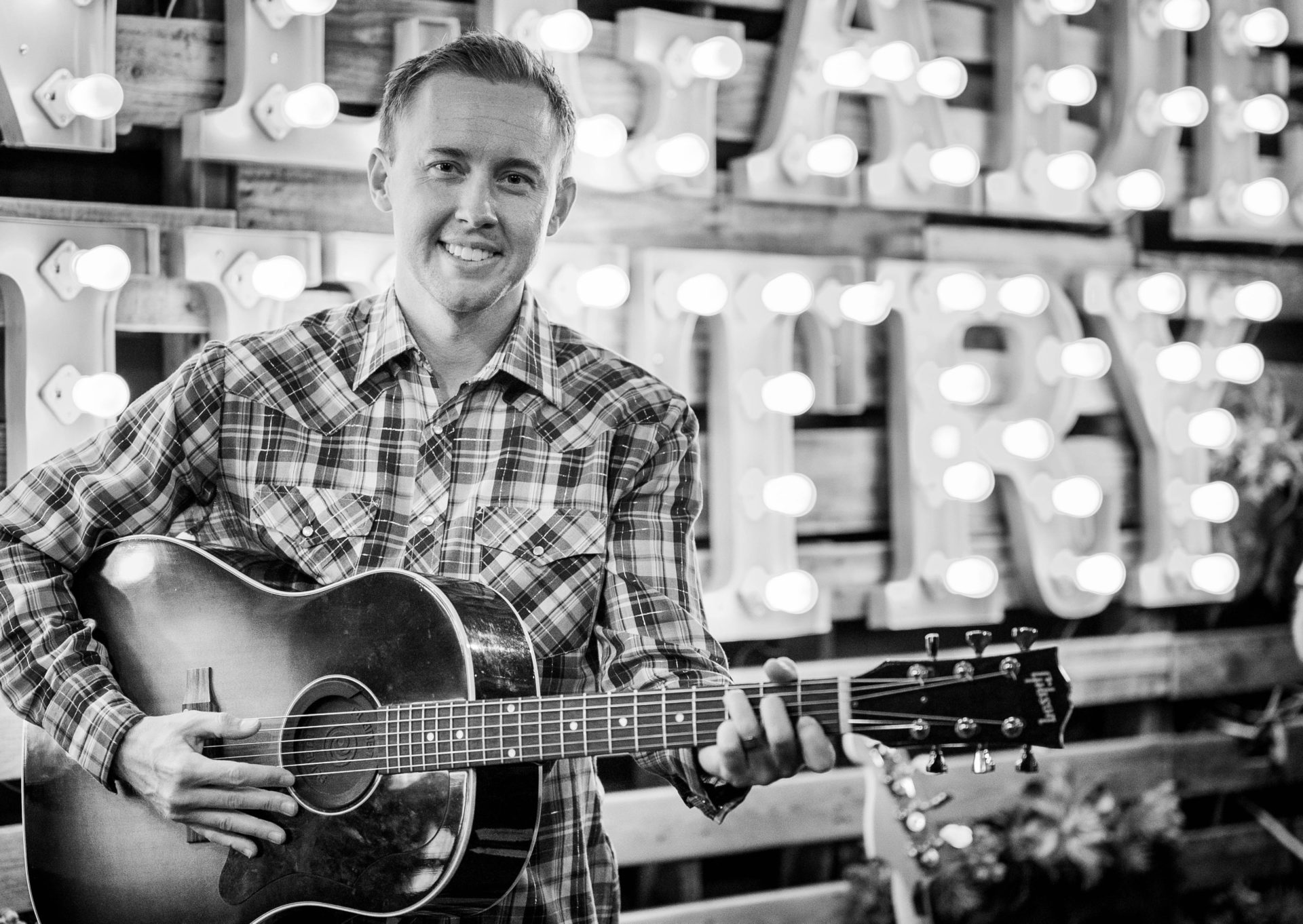 black and white photo of cody carter holding a guitar and wearing a checkered shirt standing in front of lights