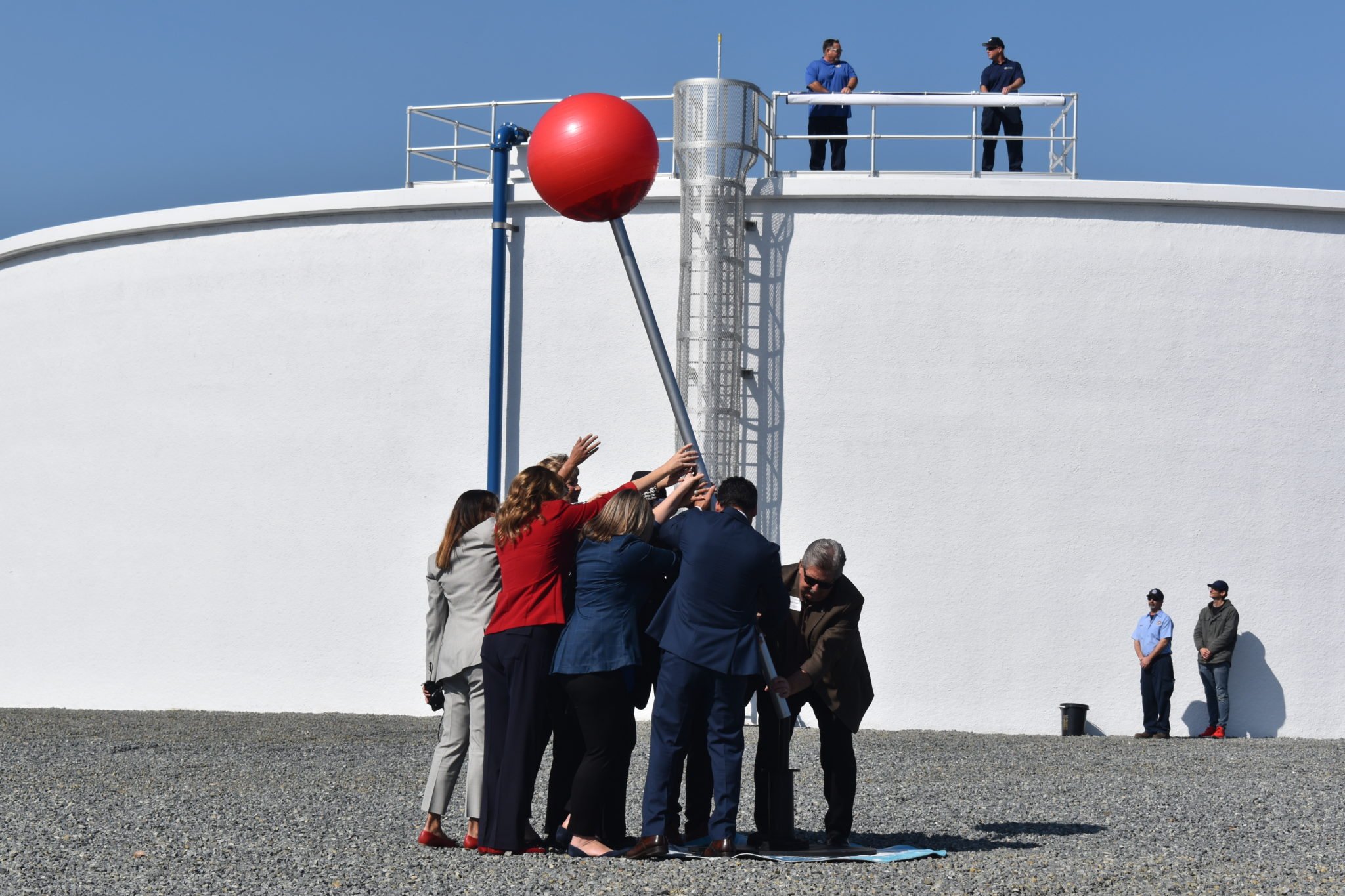 Group of people lift a red google maps location pin in front of a water plant