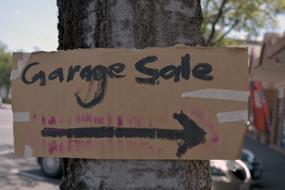 A hand made cardboard sign for a garage sale that is taped to a tree