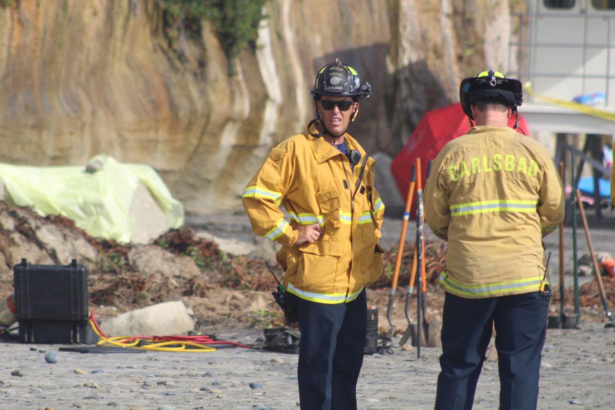 Firefighters stand in front of a large piece of sandstone that broke loose from the cliff and crashed onto Grandview Beach. File photo/The Coast News
