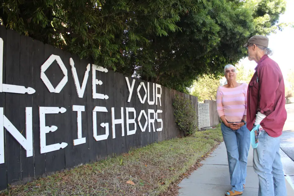 Encinitas residents JT Moring and Mary Jo Preti stand Nov. 21 at the fence where they installed a “Love Your Neighbors” message which the city ordered removed earlier this week. Photo by Jordan P. Ingram