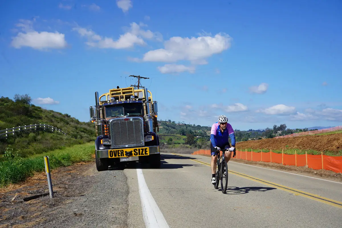 A truck hauling an oversized scraper travels along West Lilac Road in Bonsall near the proposed Ocean Breeze Ranch development as a cyclist passes by. Photo by Jordan P. Ingram