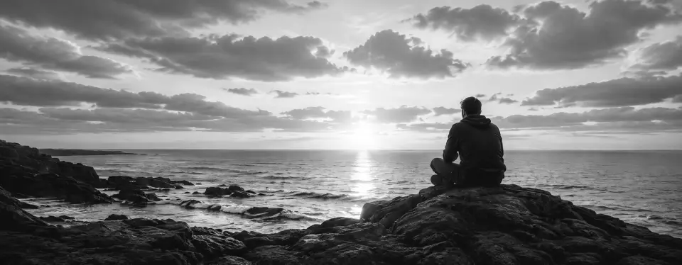 Man seated on rocky coastline overlooking the ocean at sunrise with dramatic clouds and sunlight reflecting across the water.