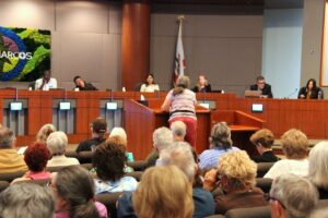 A woman speaks to people sitting on a dais in a large room.