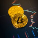 Two gold-colored Bitcoin tokens resting on a screen displaying a financial trading chart.