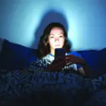 The girl lies at home in the evening on the bed with a phone in the dark.