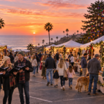 AI illustration of a sunset holiday market near Moonlight Beach, with outdoor shoppers, carolers and a Christmas tree glowing behind vendor tents.